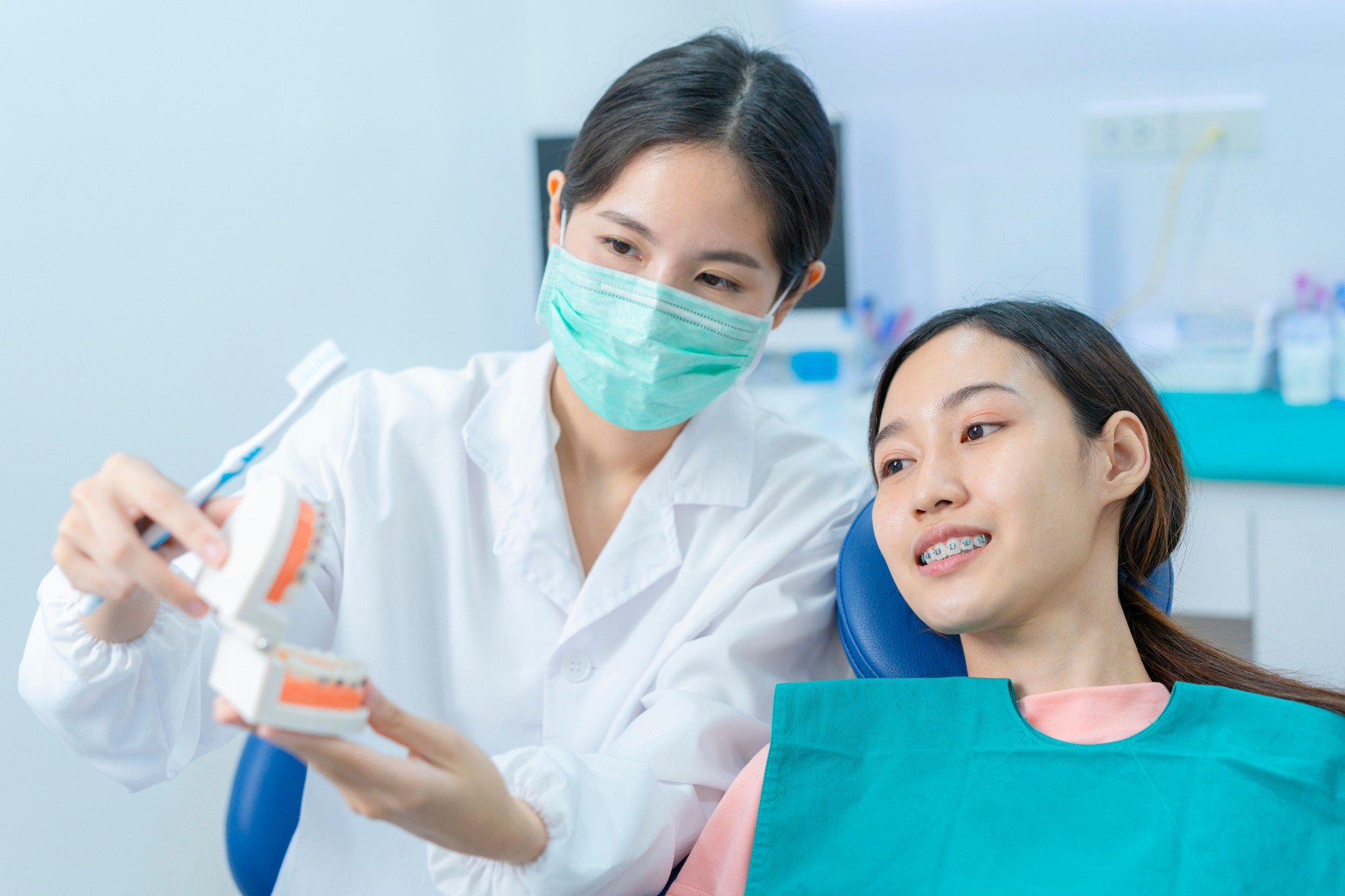 Asian young girl listening female dentist explaining about oral care and teeth brushing for people who have orthodontic. Orthodontist wearing protective face mask in dental clinic to prevent COVID-19.